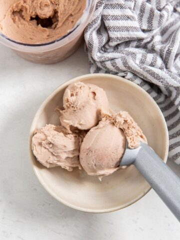 strawberry banana ice cream in a tan bowl with an ice cream scoop and a grey and white napkin