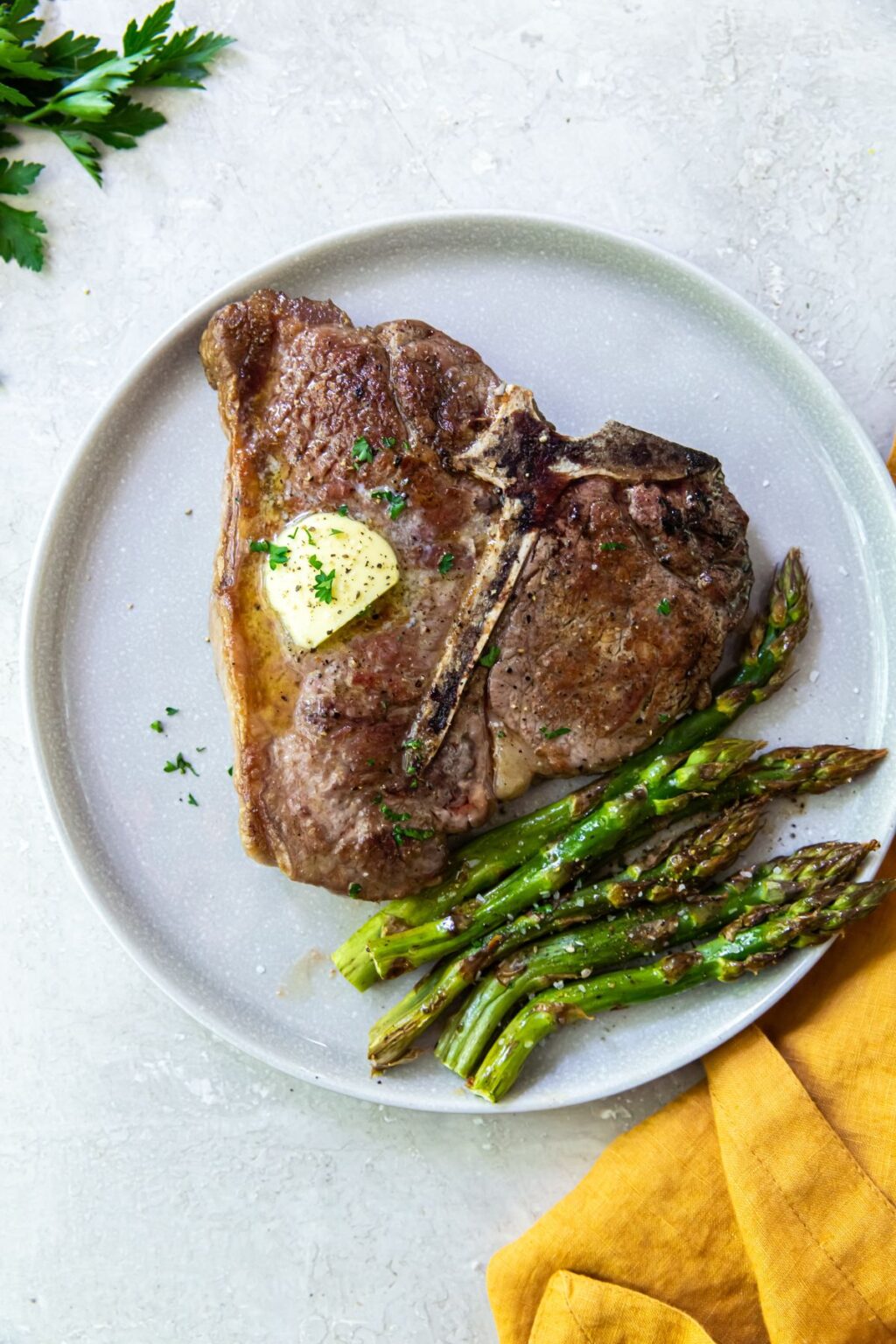 Seared Porterhouse Steak with butter, parsley, and asparagus on a white plate.