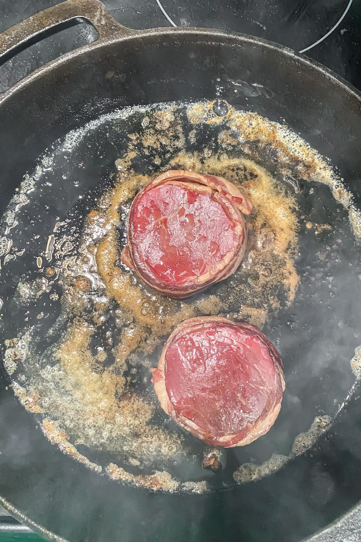 Two pieces of smoked bacon-wrapped chuck tender filets being cooked in a frying pan.