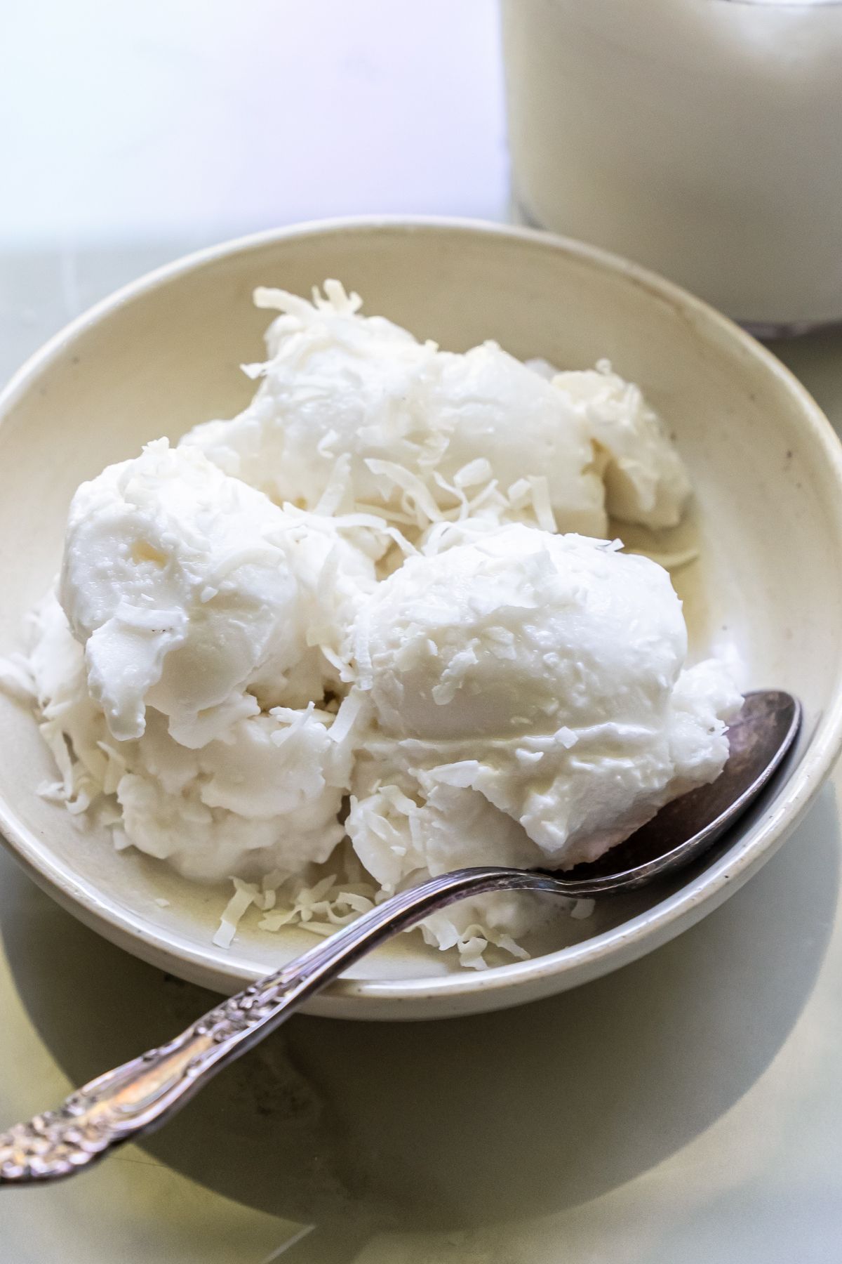 A white bowl with several scoops of white ice cream, crafted from Ninja Creami recipes, garnished with shredded coconut, and a silver spoon on the side.
