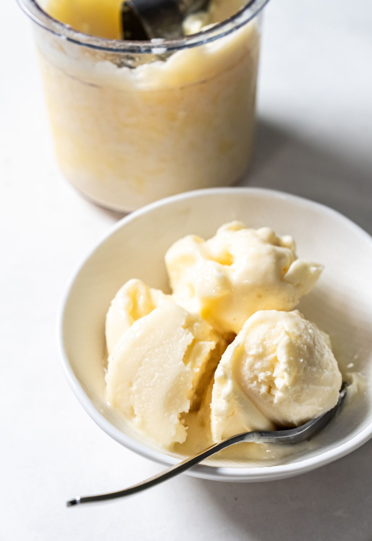 A bowl of creamy, pale yellow ice cream with a spoon. An ice cream container with similar contents is in the background, following one of the delicious Ninja Creami recipes.