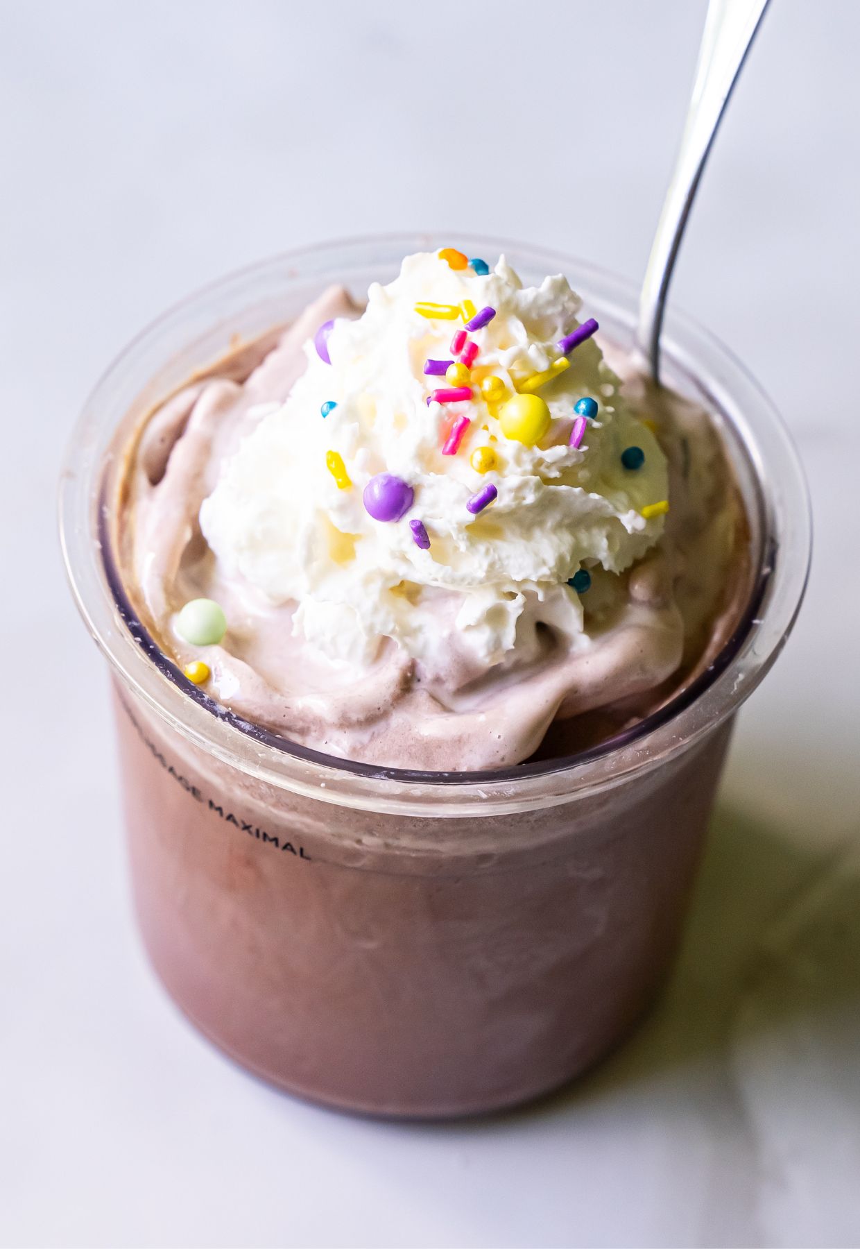 A cup of chocolate milkshake topped with whipped cream, colorful sprinkles, and a spoon inserted—a delightful treat inspired by Ninja Creami recipes.