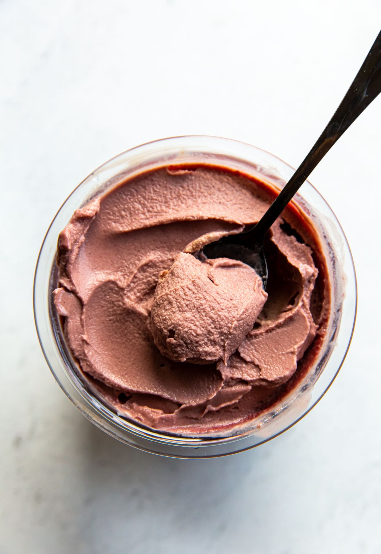 A top view of a glass jar filled with creamy chocolate cherry ice cream, prepared using Ninja Creami recipes, with a spoon scooping out a portion. The background is a light, smooth surface.