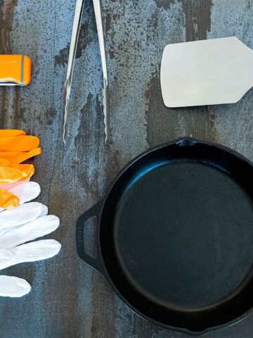 Assorted cooking utensils and protective gloves arranged around a cast iron skillet on a dark countertop.