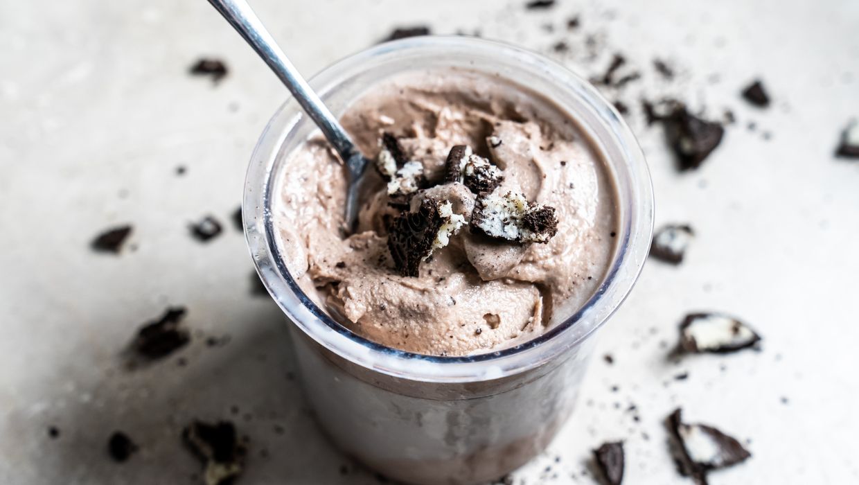 A jar of chocolate mousse topped with crumbled cookies, served with a spoon, on a speckled surface.