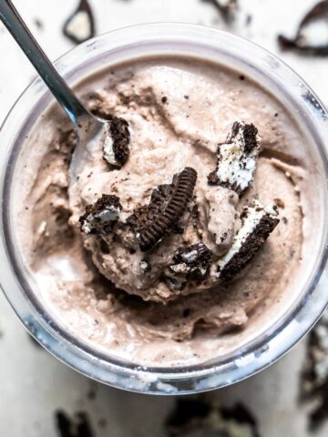 Top view of a glass of chocolate ice cream with crushed cookies, served with a spoon on a speckled surface.