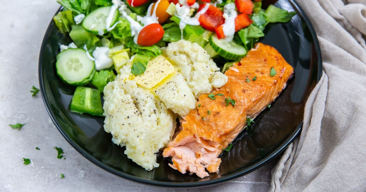 A plate containing a piece of cooked salmon, a serving of mashed potatoes with butter, and a salad with cucumbers, tomatoes, red bell peppers, and a drizzle of dressing.