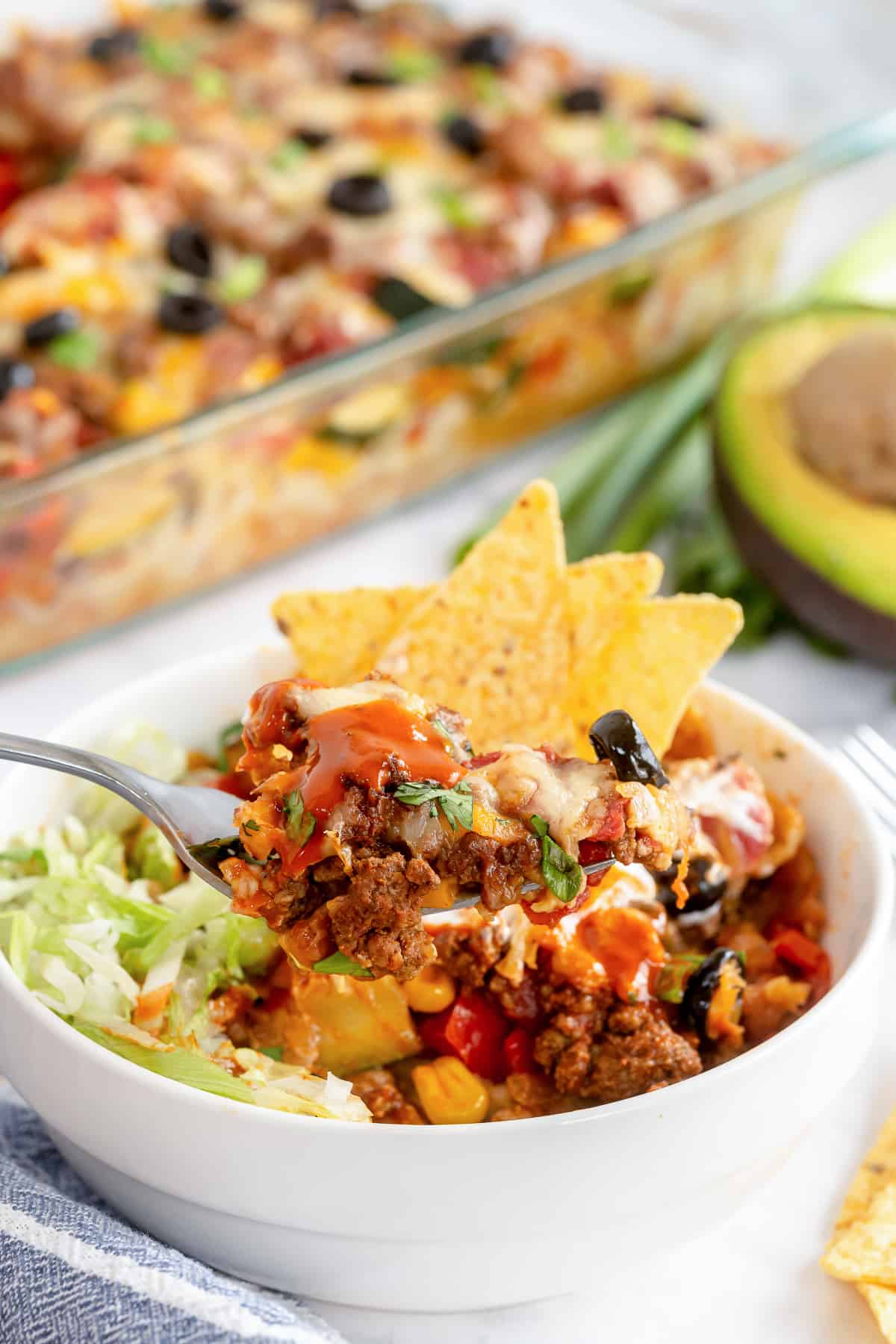 A bowl of taco casserole topped with tortilla chips and sauce, with a spoon lifting a bite. In the background, another casserole dish brims with the same savory delight, and a half avocado is visible beside the bowl.