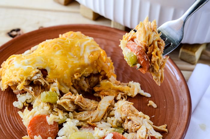 A close-up of a forkful of casserole with shredded chicken, vegetables, and melted cheese, lifted from a red plate that holds more casseroles of the dish.