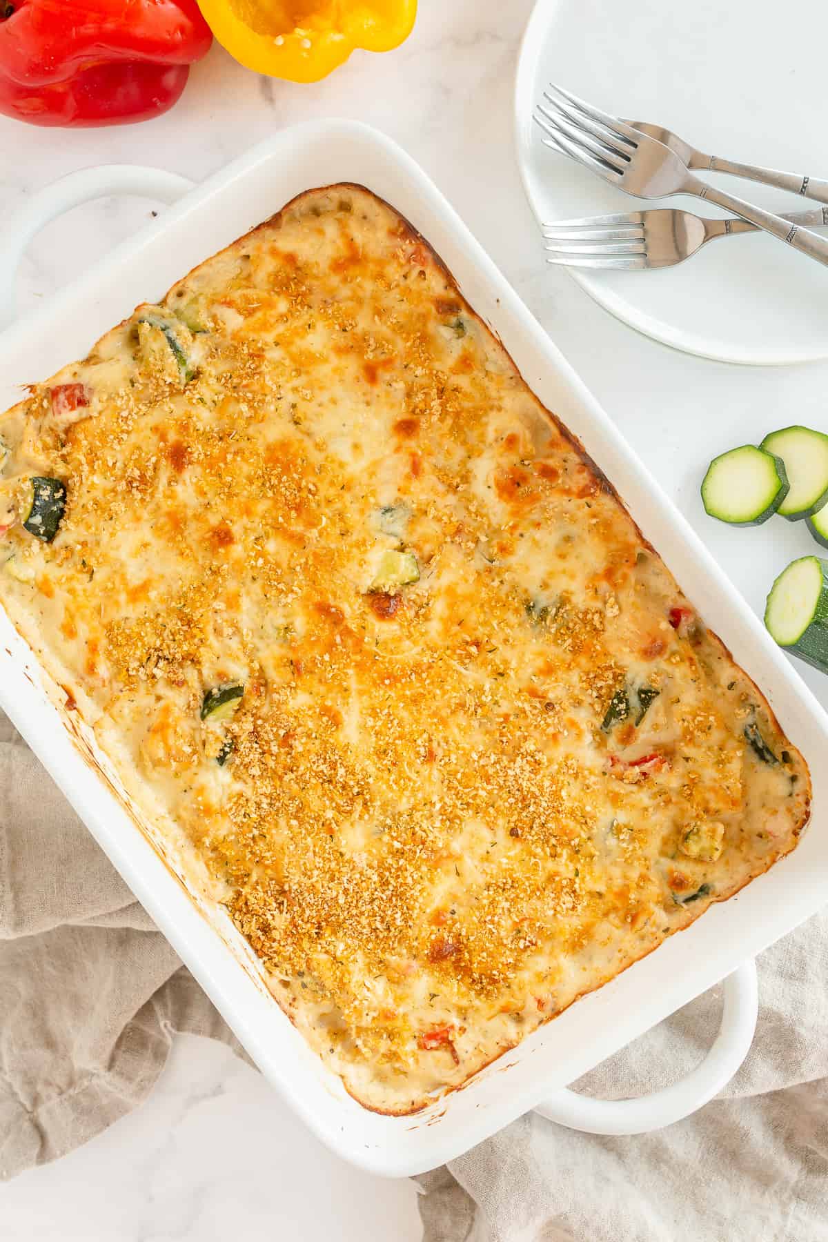 A rectangular baking dish with a baked casserole topped with a golden-brown crust. Sliced zucchini and bell peppers are beside it, showcasing the versatility of casseroles. Plates and forks are set nearby on a white surface.