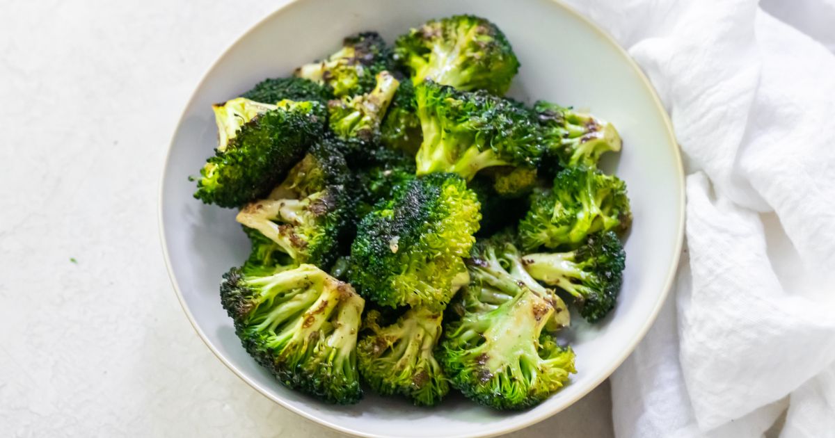 A bowl of roasted broccoli florets on a white table, accompanied by a white napkin.