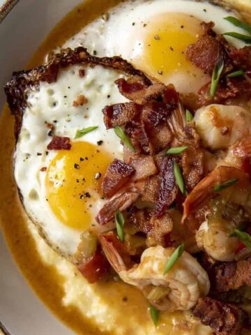 A bowl of creamy grits topped with two sunny-side-up eggs, shrimp, crumbled bacon, chopped green onions, and a drizzle of sauce made effortlessly in an Instant Pot.