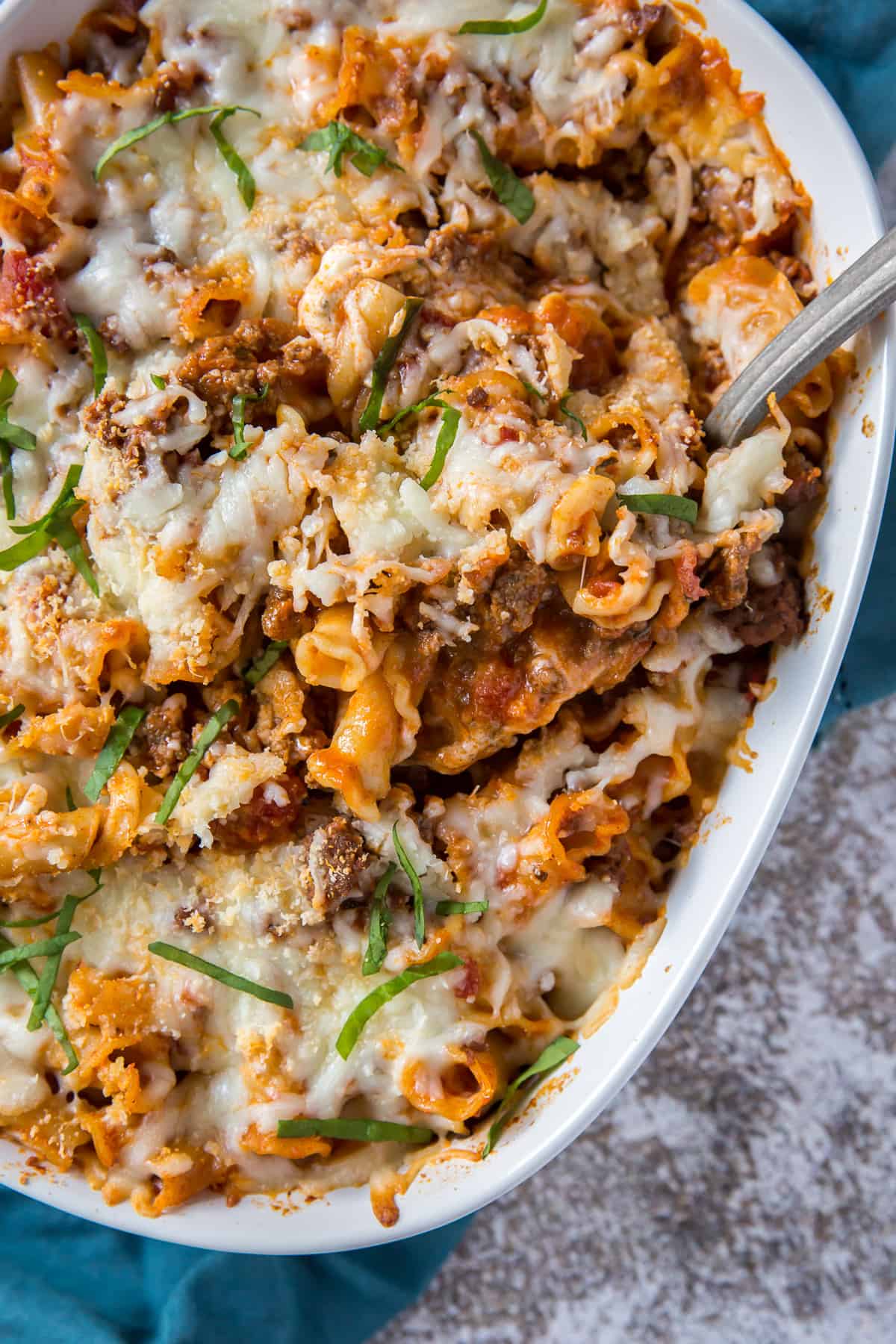 Baked pasta casserole with melted cheese, ground beef, and garnished with chopped fresh basil in a white ceramic baking dish. A serving spoon is placed in the dish.