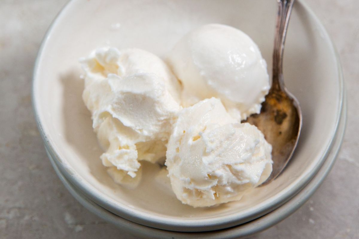 Bowl of vanilla ice cream with a spoon, partially eaten, on a light gray surface.