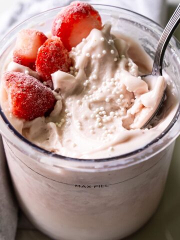 A glass filled with creamy yogurt topped with fresh strawberries and small white sprinkles, with a spoon in it. A grey napkin lies next to the glass.