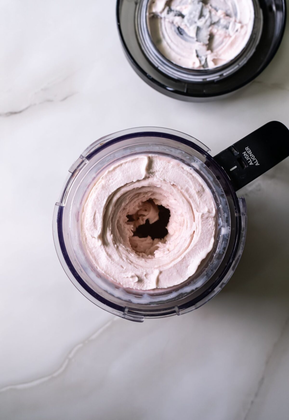 A top view of a food processor bowl filled with creamy, light pink mixture and an attached lid with a black handle placed nearby on a marble surface.