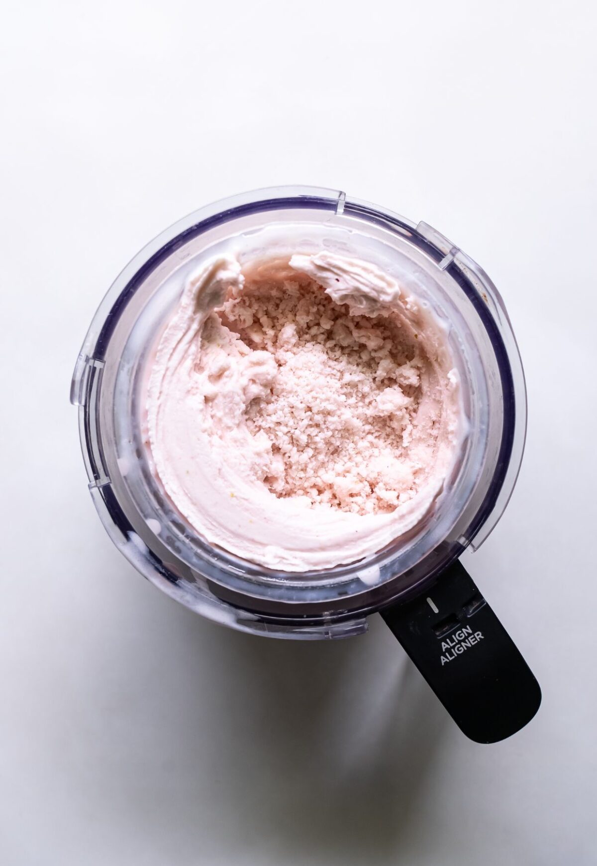 A top-down view of a pale pink creamy substance in a food processor against a white background.