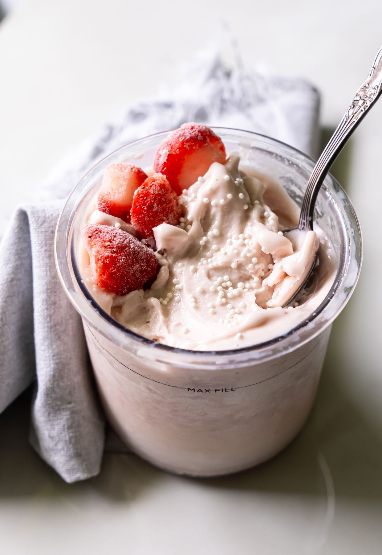 A glass jar filled with creamy yogurt topped with whole strawberries and white sprinkles, with a metal spoon inside, placed on a light surface with a gray cloth napkin beside it.