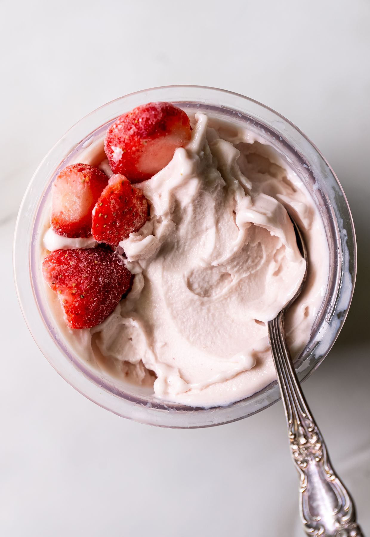 A glass container filled with whipped cream topped with sliced strawberries. A silver spoon is placed inside the container.
