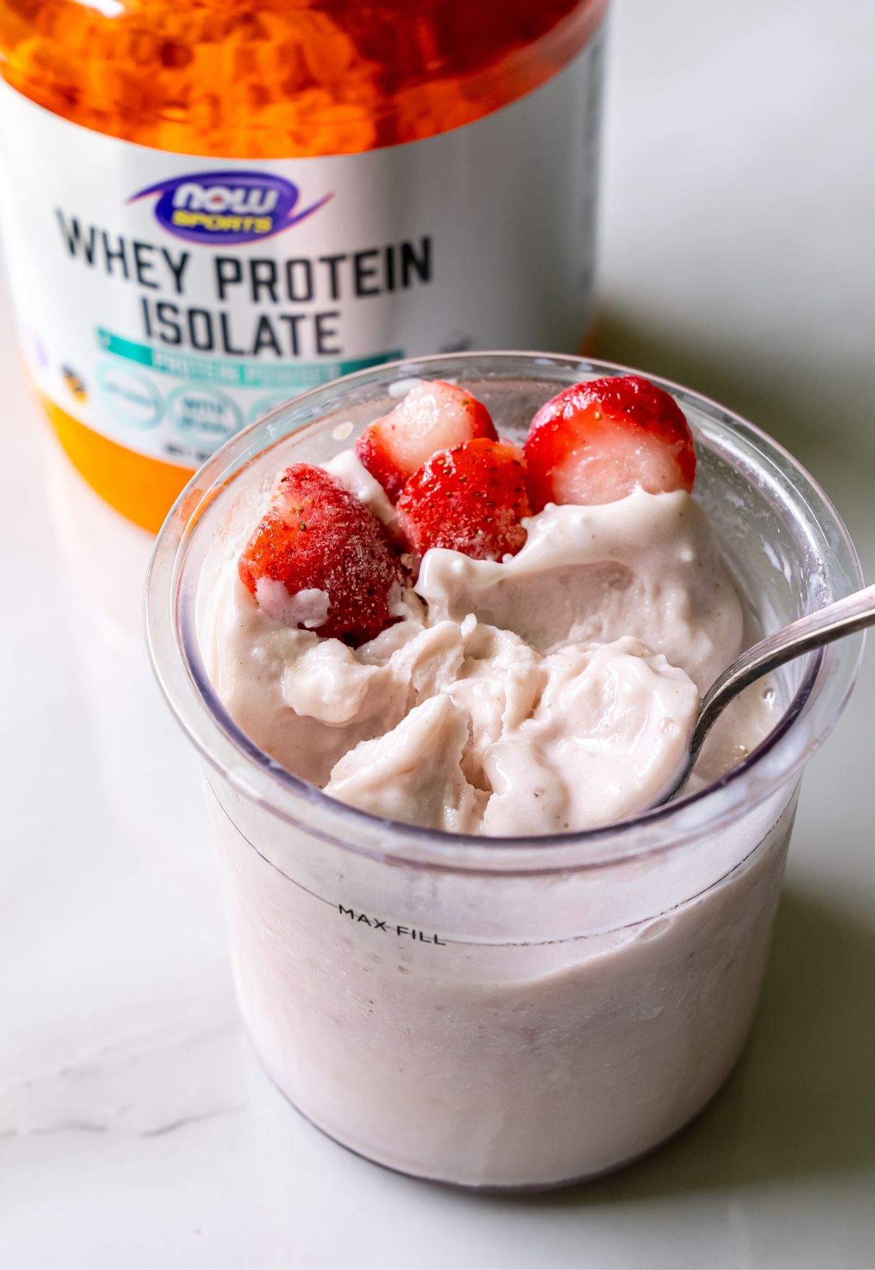 A container of whey protein isolate with a bowl of creamy yogurt topped with sliced strawberries and a spoon.