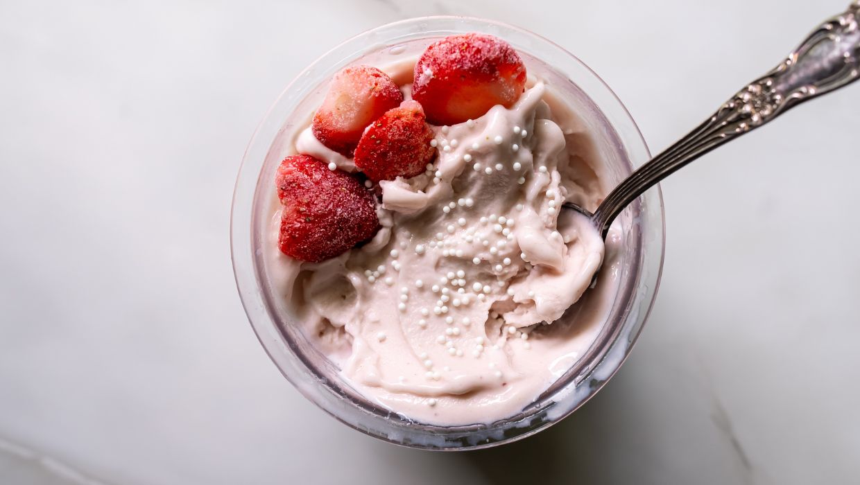 A glass bowl filled with strawberry ice cream topped with frozen strawberries and small white sprinkles, with a metal spoon inserted on the right side.