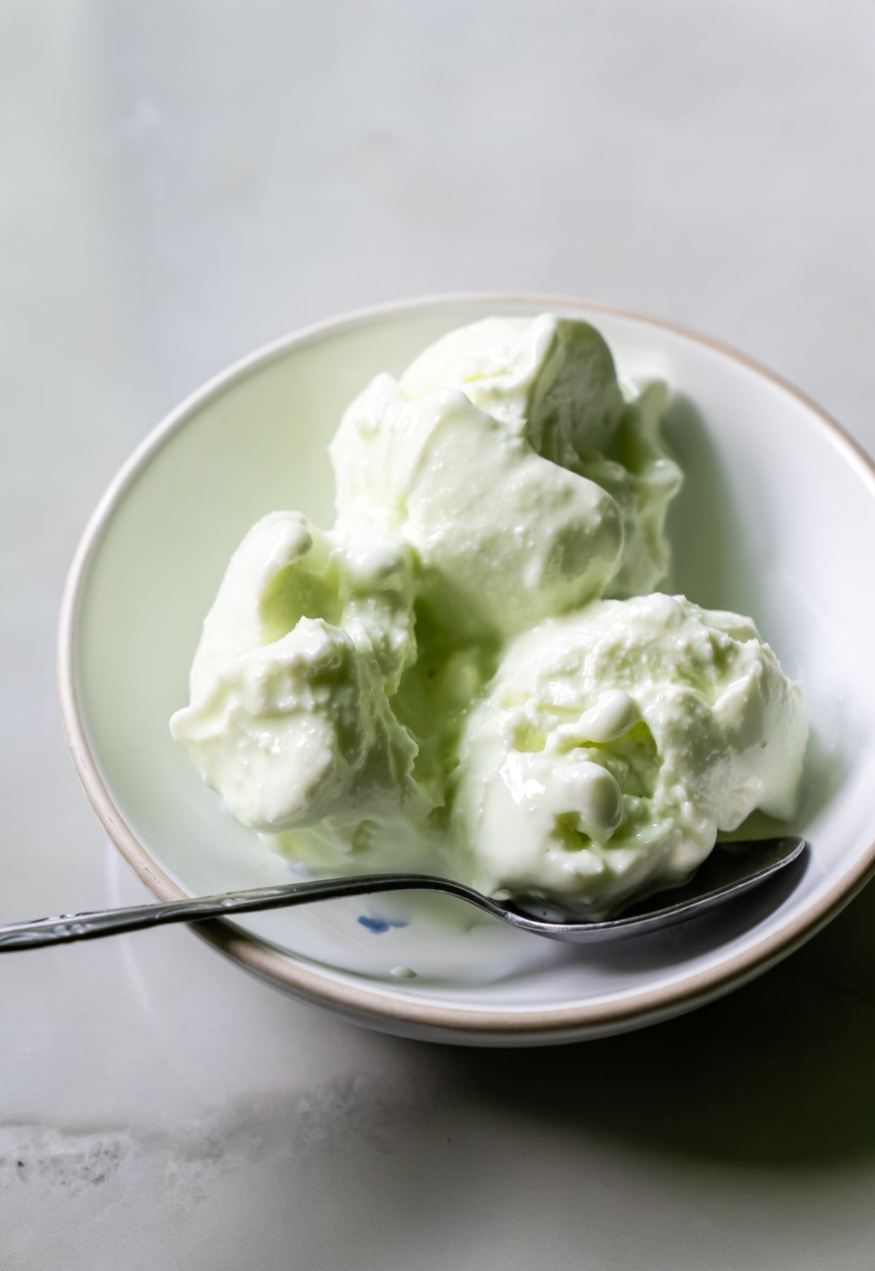 A white bowl containing three scoops of green ice cream, a delightful dessert, with a metal spoon placed beside them on a light gray surface.