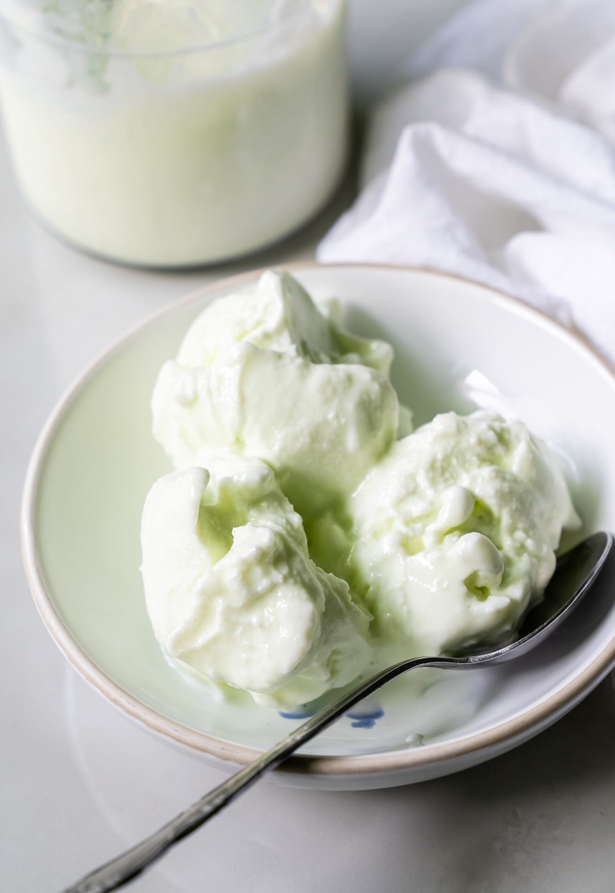 A bowl with three scoops of light green ice cream, an irresistible sight among desserts, and a spoon rests on a white surface. A jar with more ice cream and a white cloth are visible in the background.