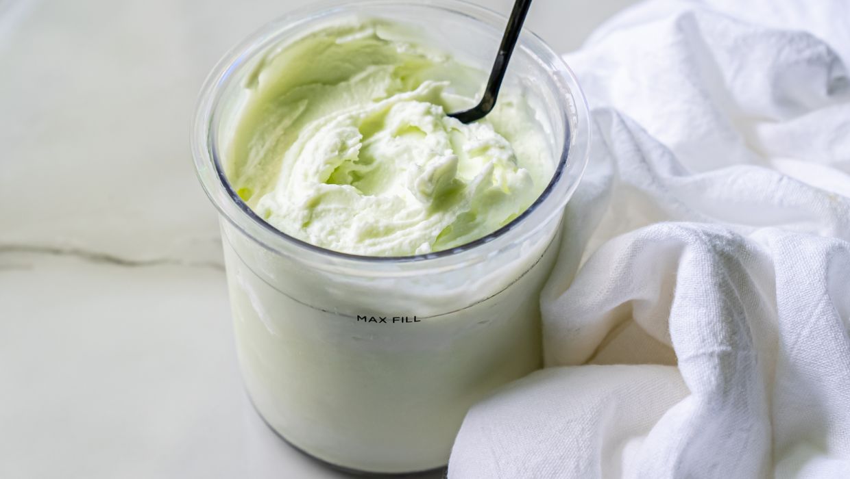 A clear container filled with a light green, creamy dessert is complemented by a black spoon inside. A white cloth is neatly placed next to the container.