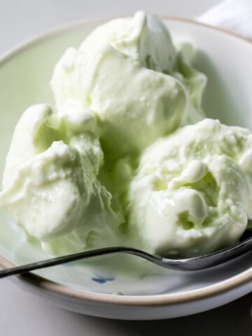 A white bowl contains three scoops of light green ice cream, a delightful dessert with a spoon resting on the bowl's edge. A white cloth is partially visible next to the bowl.