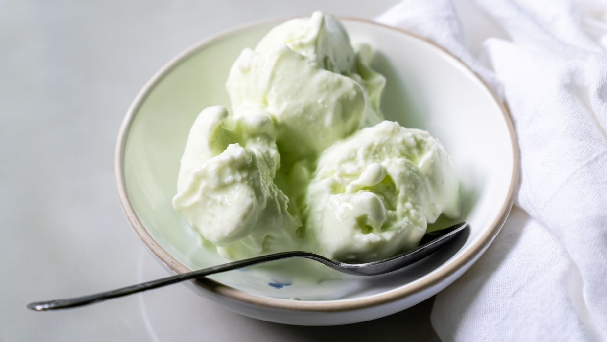 A white bowl contains three scoops of light green ice cream, a delightful dessert with a spoon resting on the bowl's edge. A white cloth is partially visible next to the bowl.