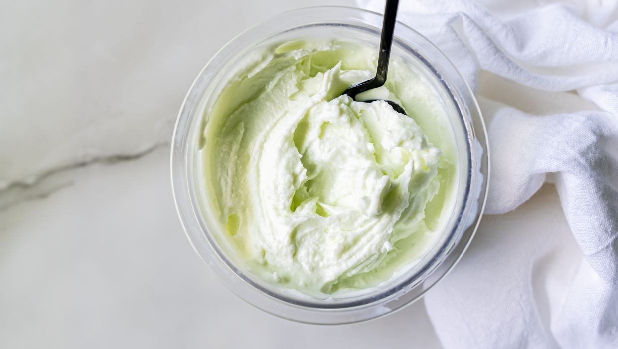 A clear plastic container of pale green ice cream with a black spoon sticking out, placed next to a white cloth on a marble surface, showcases an inviting dessert scene.