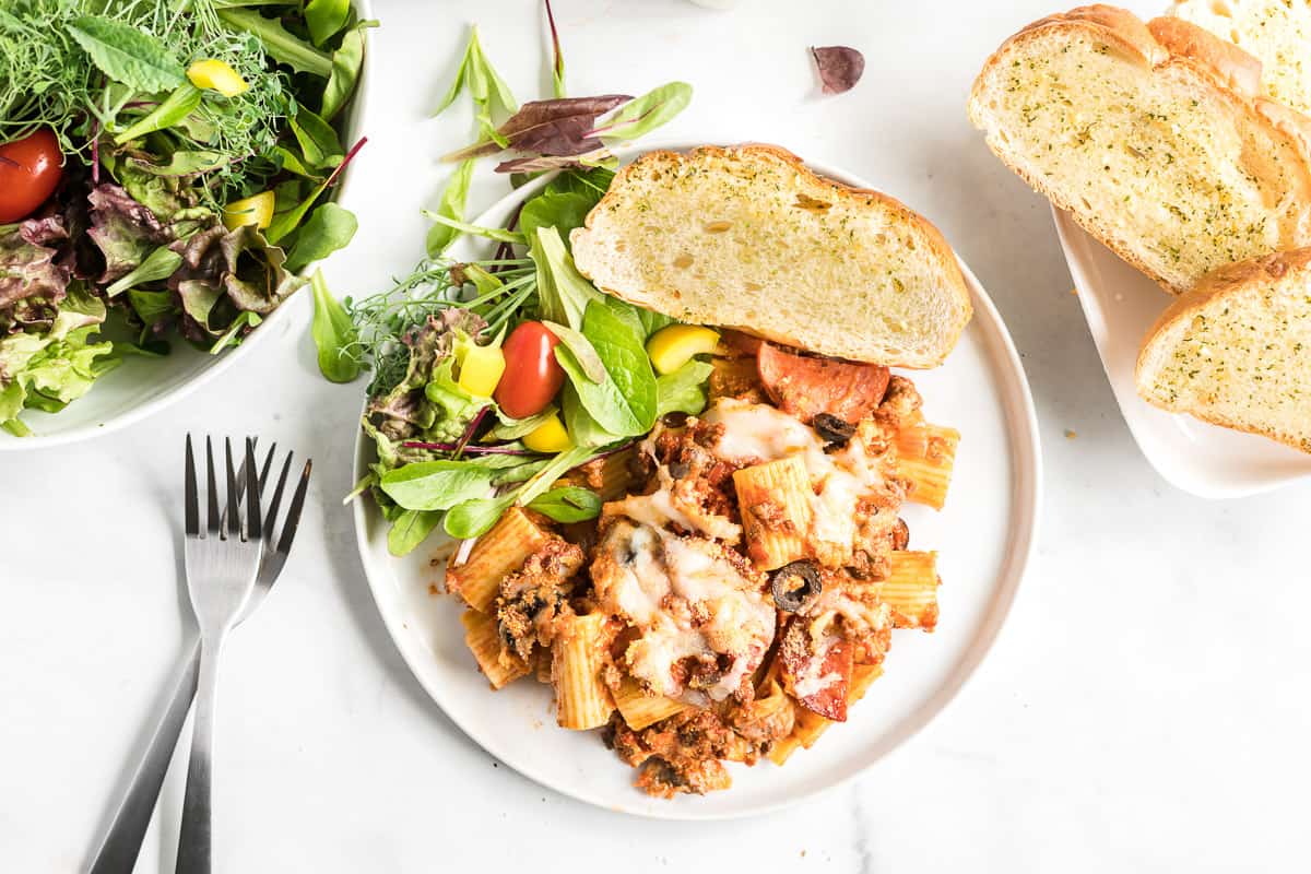 A plate with baked pasta, reminiscent of comforting casseroles, topped with melted cheese, served alongside a mixed salad with cherry tomatoes and two slices of garlic bread. Forks and a bowl of salad are beside the plate.