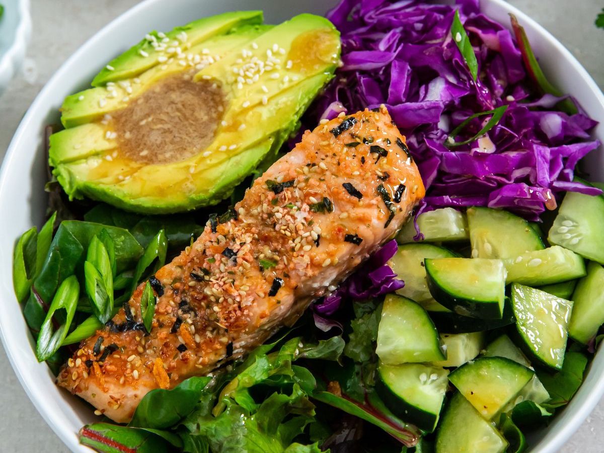 A dinner bowl containing a portion of grilled salmon, sliced avocado sprinkled with sesame seeds, chopped purple cabbage, fresh cucumber pieces, and leafy greens.