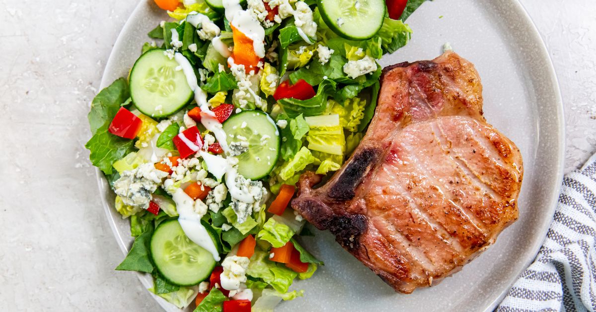 A grilled pork chop next to a mixed green salad with cucumbers, bell peppers, blue cheese, and dressing on a white plate.