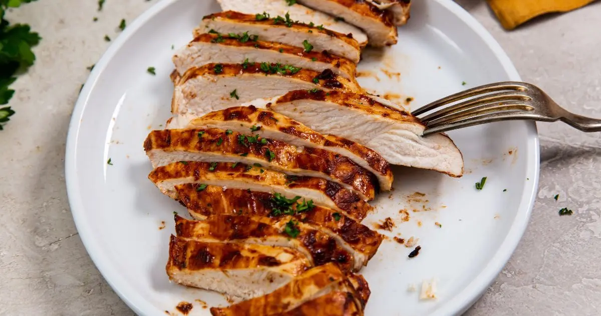 Sliced grilled chicken breast on a white plate garnished with herbs, with a fork on the side.