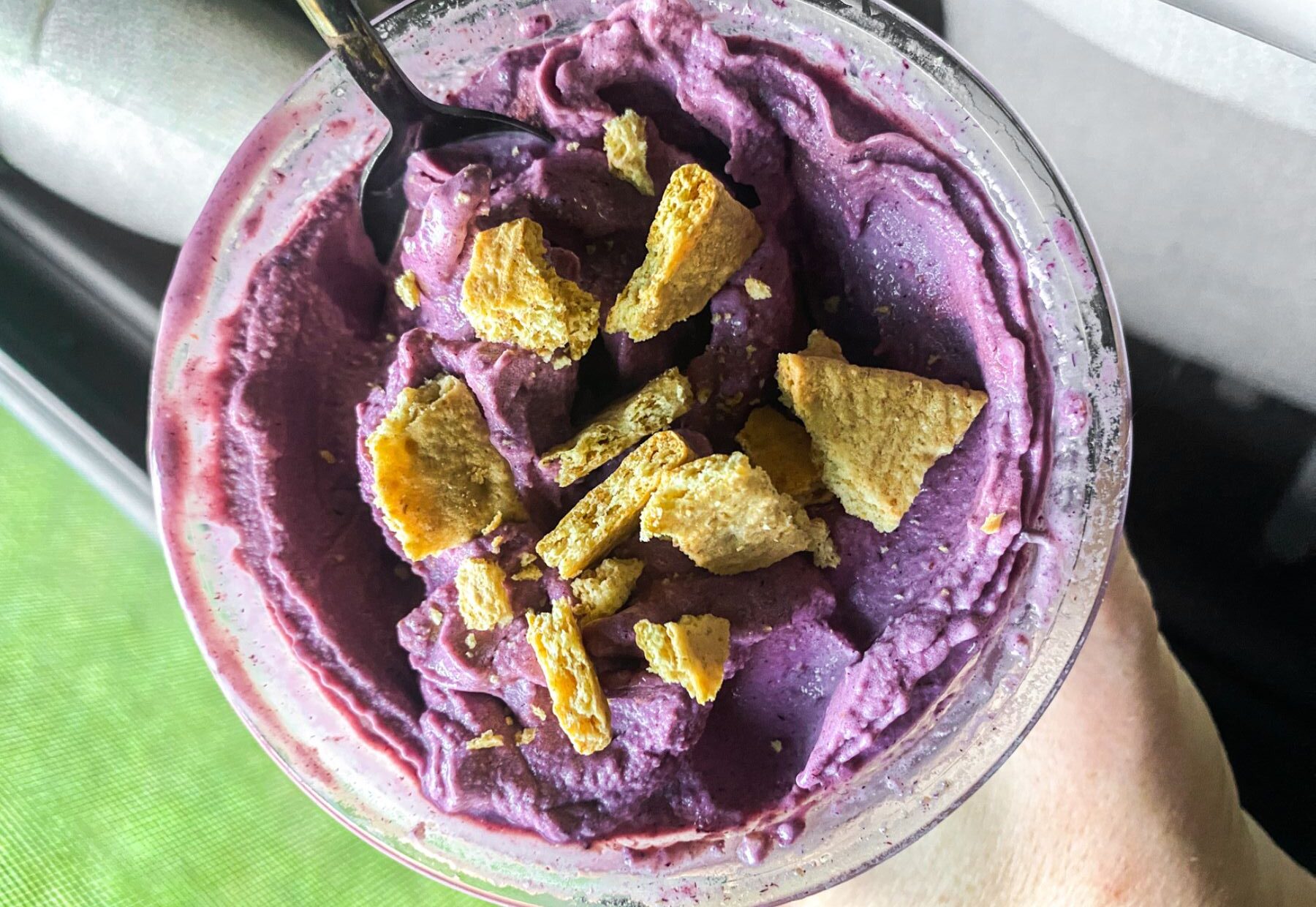 A glass cup filled with purple acai smoothie topped with crushed pieces of cereal, with a black spoon in it.