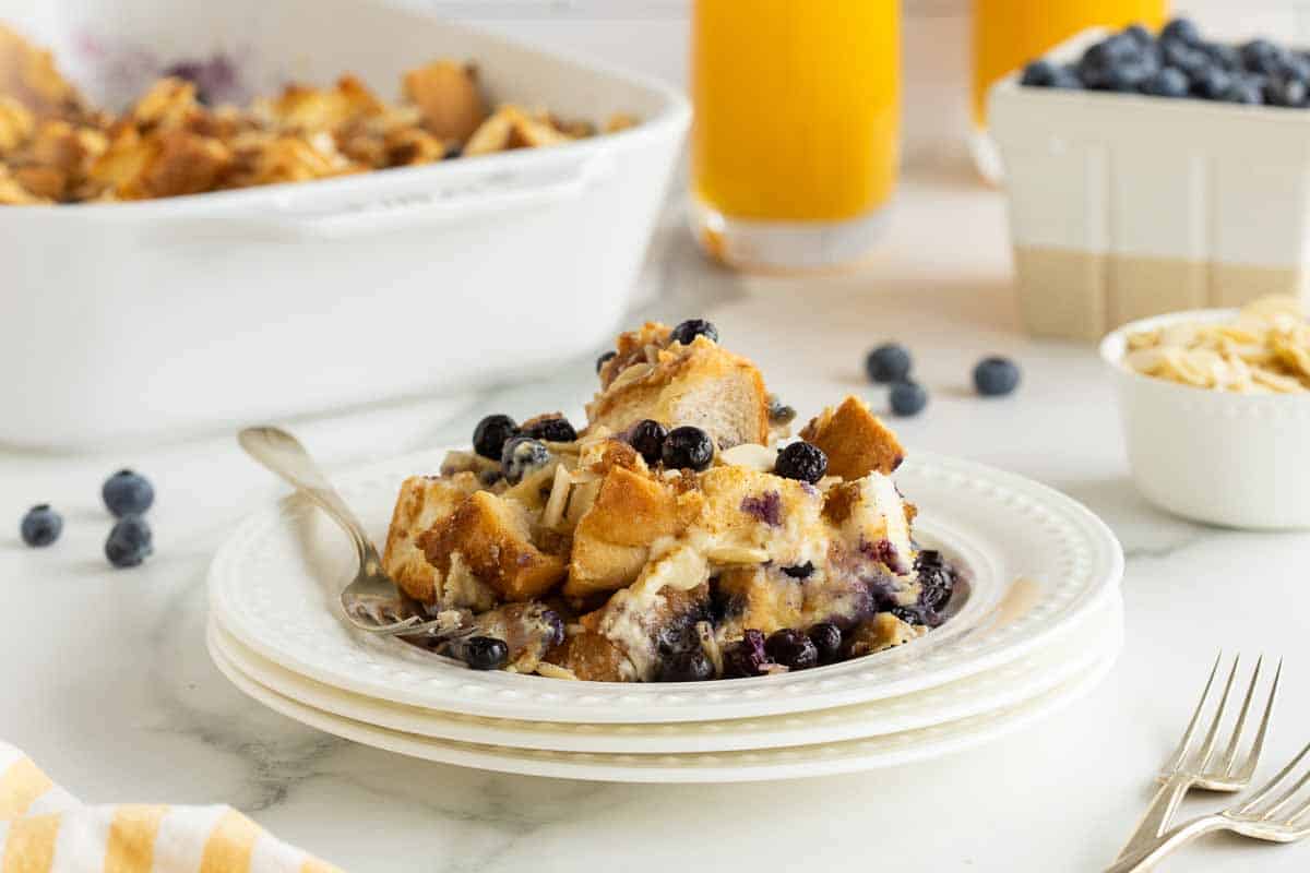 A plate of blueberry breakfast casserole placed on a white plate with a fork. In the background, there is a dish of casseroles, glasses of orange juice, and a container of fresh blueberries.