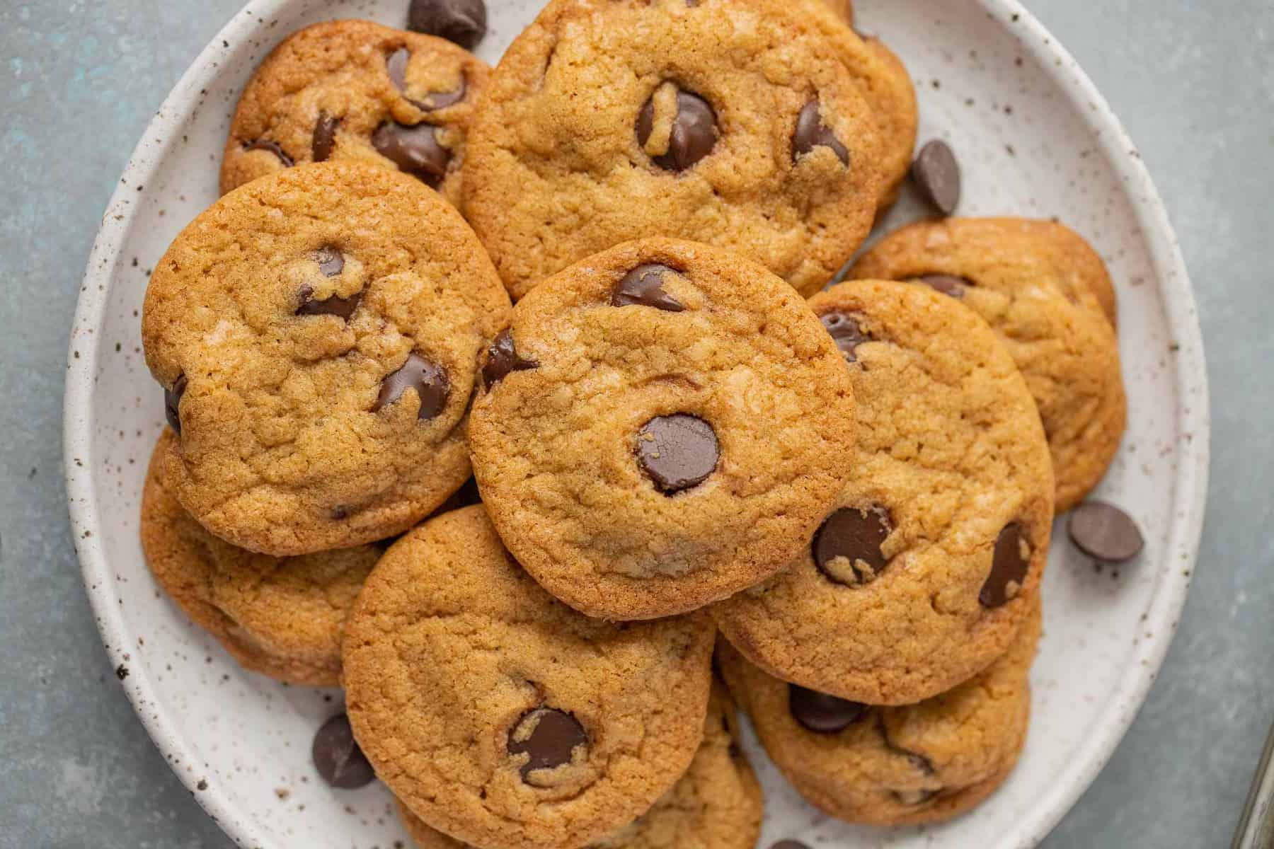 A plate of freshly baked chocolate chip cookies with visible chocolate chunks, displayed on a light grey surface.