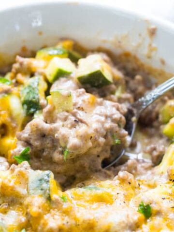 A close-up of a bowl of cheesy ground beef and vegetable casserole, featuring zucchini chunks and garnished with chopped herbs, showcases one of the heartiest casseroles around. A spoon rests invitingly in the dish.