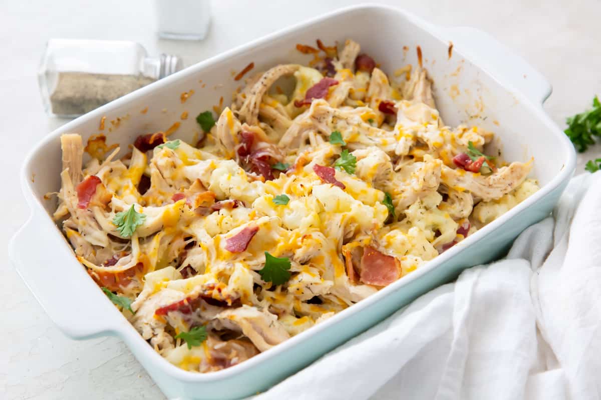 Keto Chicken Bacon Ranch with cauliflower, cheese, and parsley in a white casserole dish with a wooden spoon.