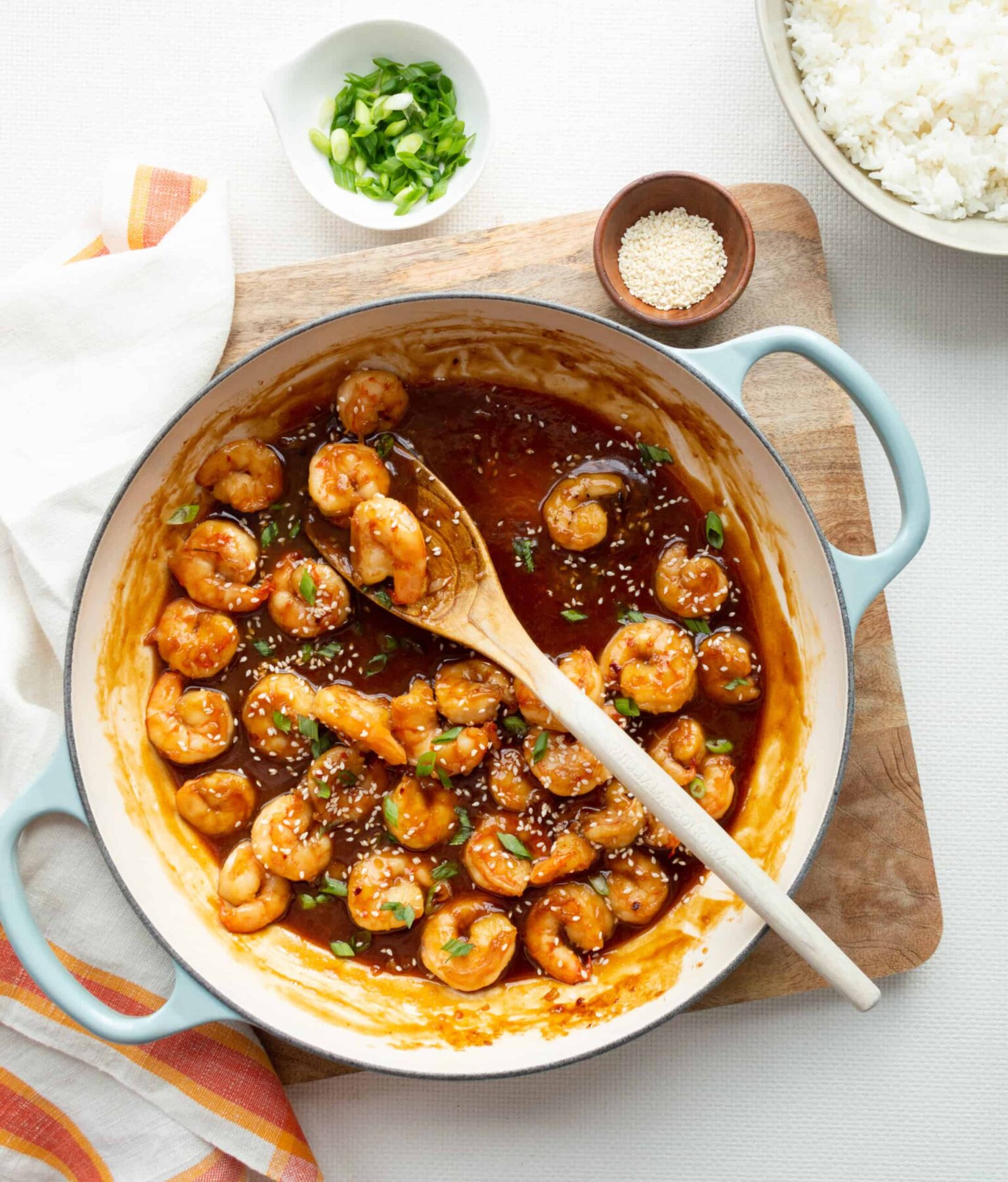 A wooden spoon in an aqua handled cast-iron pan with honey-garlic shrimp on top of a wooden board.