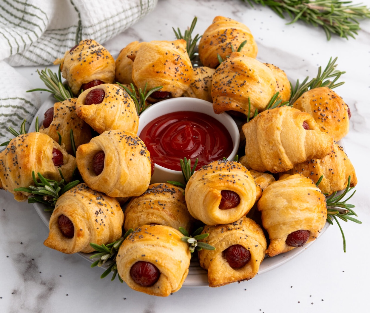 A tempting plate of pastry-wrapped sausages, sprinkled with poppy seeds, serves as savory appetizers, arranged around a dish of ketchup and garnished with sprigs of rosemary on a sleek marble surface.