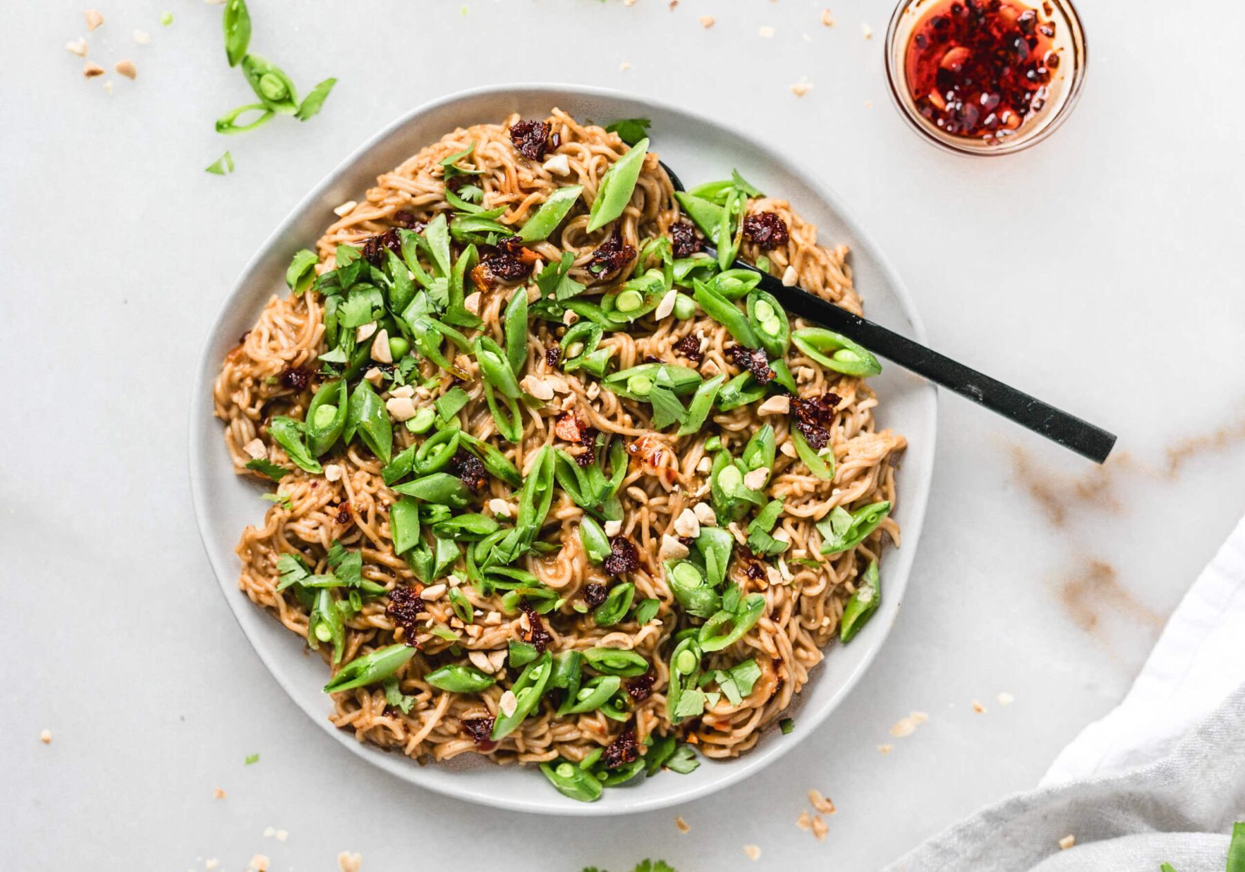 A plate of noodles mixed with green vegetables and garnished with herbs sits on a white surface, perfect for dinners. A black chopstick rests on the plate's edge, while a small jar of red chili oil is adjacent to the plate.