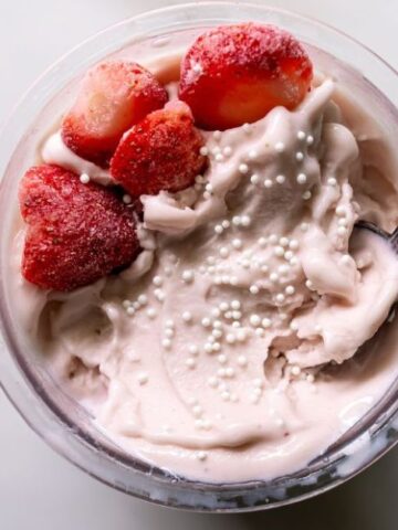 A bowl of strawberry yogurt topped with whole strawberries and white sprinkles, with a spoon inside.