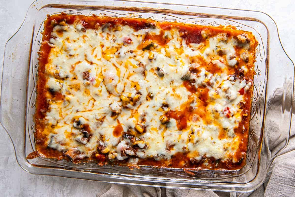A glass baking dish filled with a mouthwatering casserole topped with melted cheese, savory meat, and rich tomato sauce rests on a light-colored surface with a beige cloth nearby.