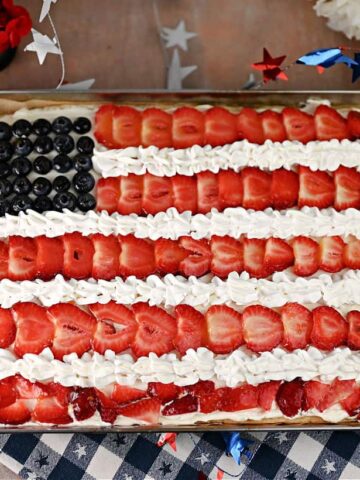 A rectangular cake decorated to resemble the U.S. flag, with blueberries as the blue field, and strawberries and whipped cream forming the red and white stripes. Red and white flowers are in the background.