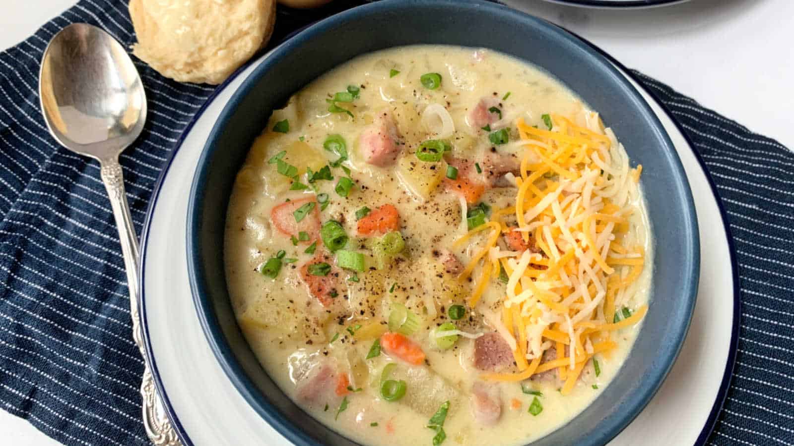 A bowl of potato soup with cheese and biscuits on a table.