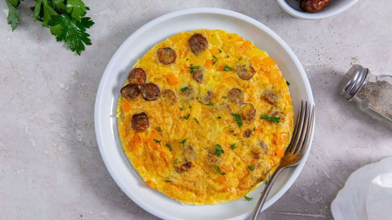 A cooked omelette with slices of sausage and chopped herbs on a white plate. A fork, parsley, a salt shaker, and a small bowl of dates are on the side on a light-colored surface.