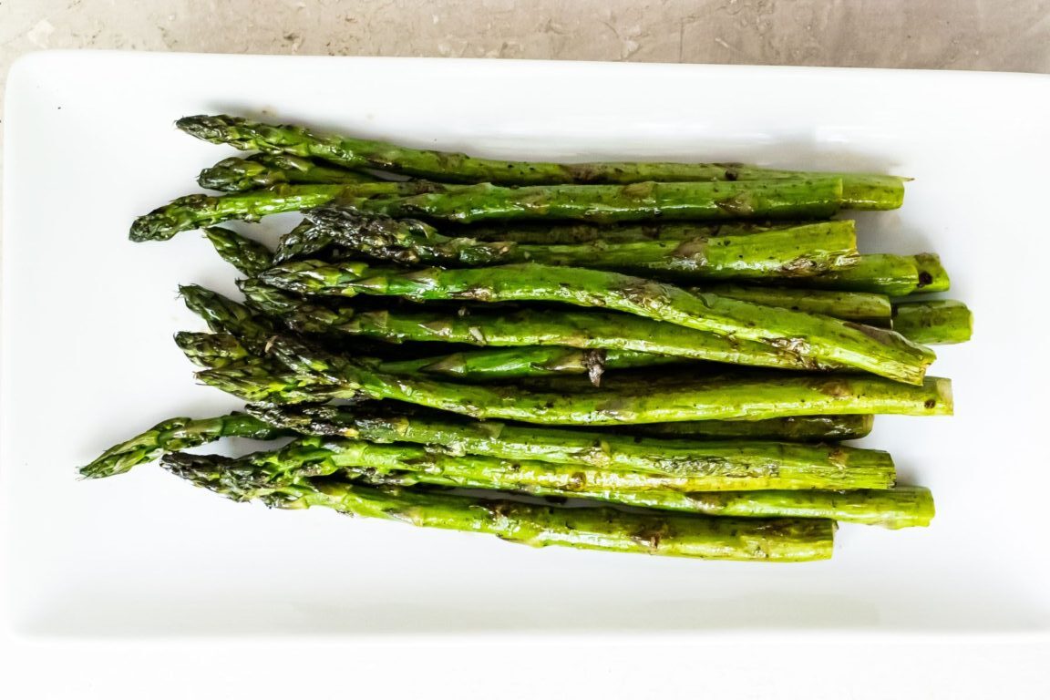 A plate of roasted asparagus spears, arranged neatly on a rectangular white dish. The asparagus has a lightly charred appearance, suggesting it has been cooked with a bit of oil or seasoning.