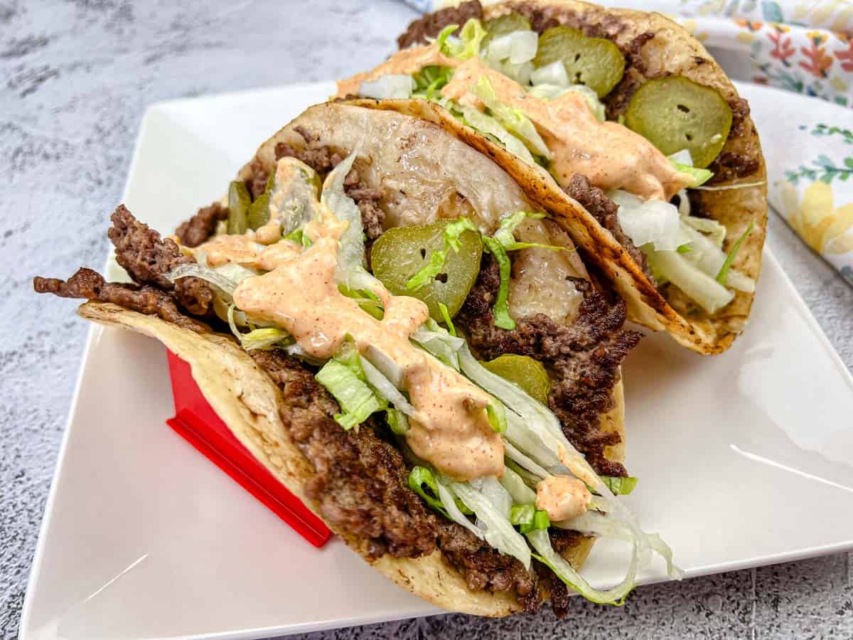 Two folded tacos filled with ground beef, shredded lettuce, pickles, and a dollop of spicy sauce, placed on a white square plate.